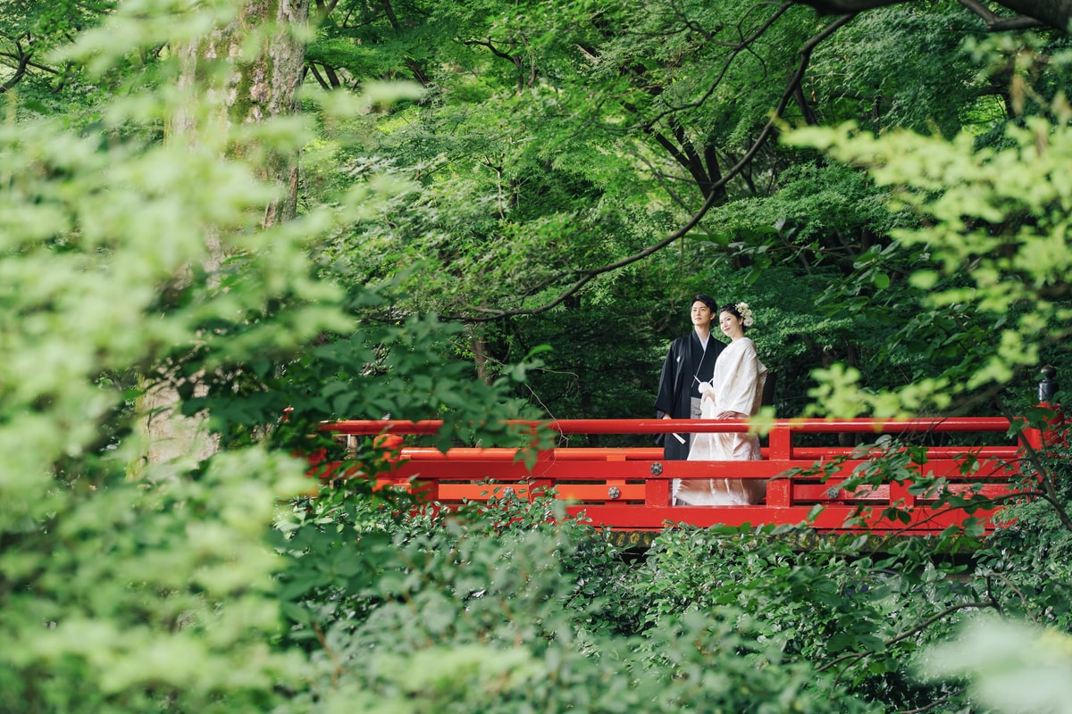 A couple in Japanese wedding attire standing on a red bridge surrounded by greenery